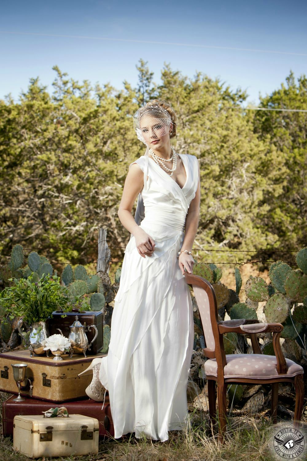 beautiful blond female model with a white flowing dress with pearls and an up-do with a birdcage veil and lace shoes stand next to vintage  chair and antique suitcases with tea set in front of cactus and barbed wire with trees and blue sky in the background